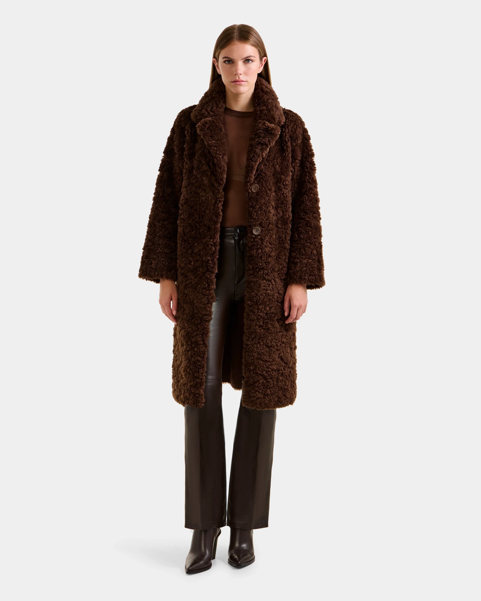 A woman faces forward wearing the Rudsak ANNABELLE FSU - L125512 Espresso sherpa teddy coat—textured, knee-length, in dark brown—layered over a brown top, dark pants, and ankle boots against a plain light background.