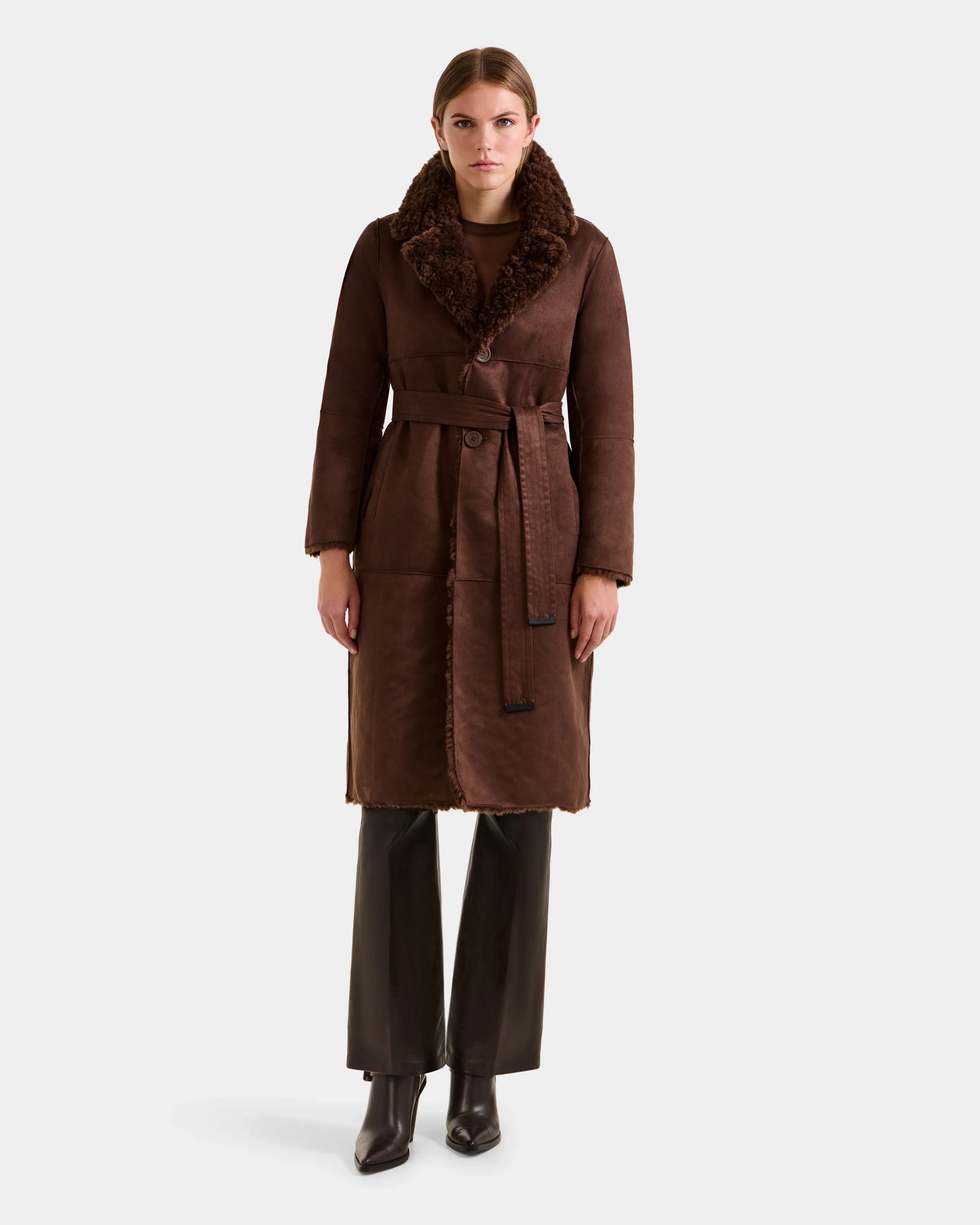 A woman faces forward against a plain light background, wearing the Rudsak ANNABELLE FSU - L125512 Espresso long faux suede trench coat with a fur collar and belt, paired with dark pants and heeled boots.
