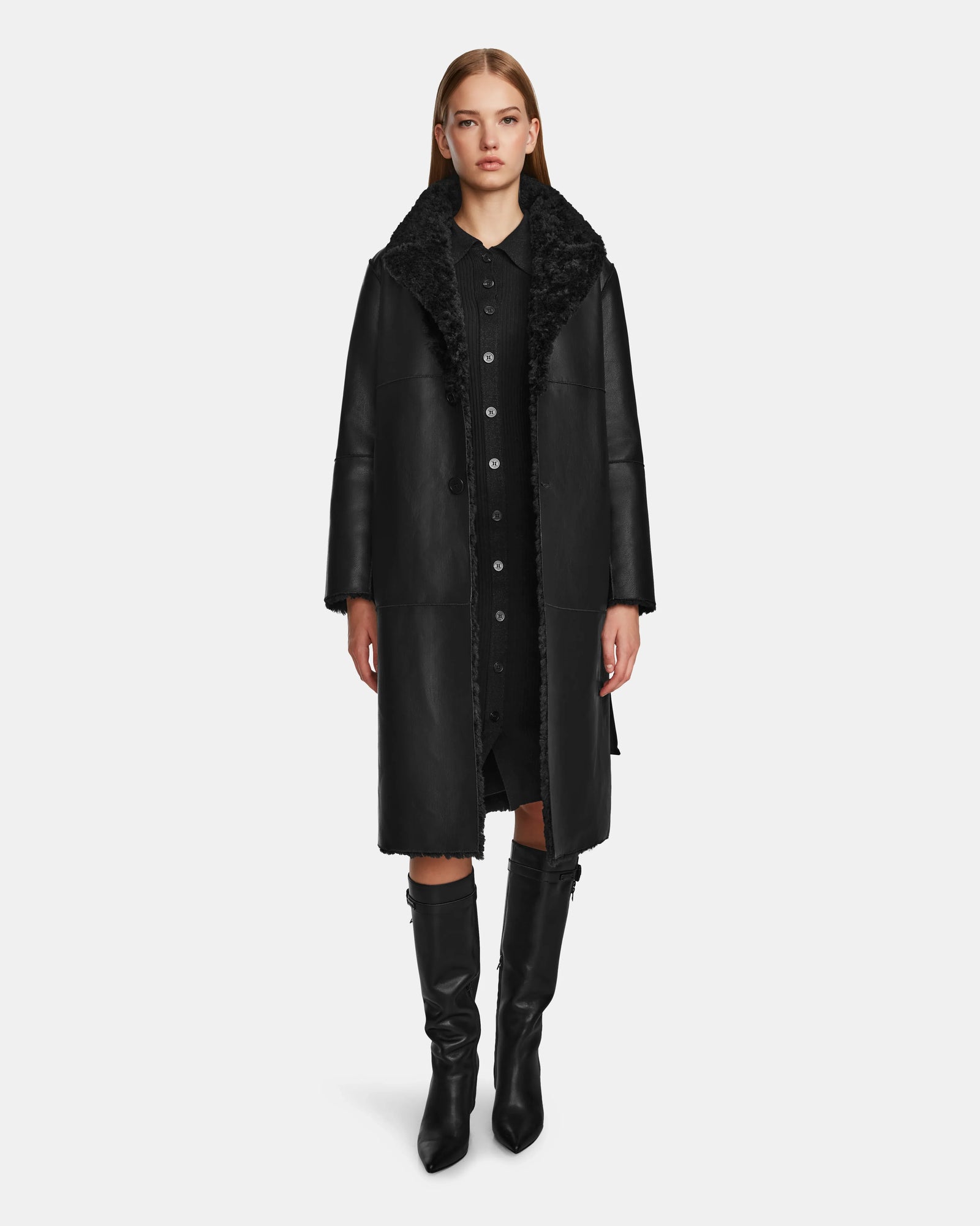 A woman faces forward, wearing the Rudsak ANNABELLE - L124544 Black vegan leather coat over a black dress and knee-high boots, set against a plain white background.