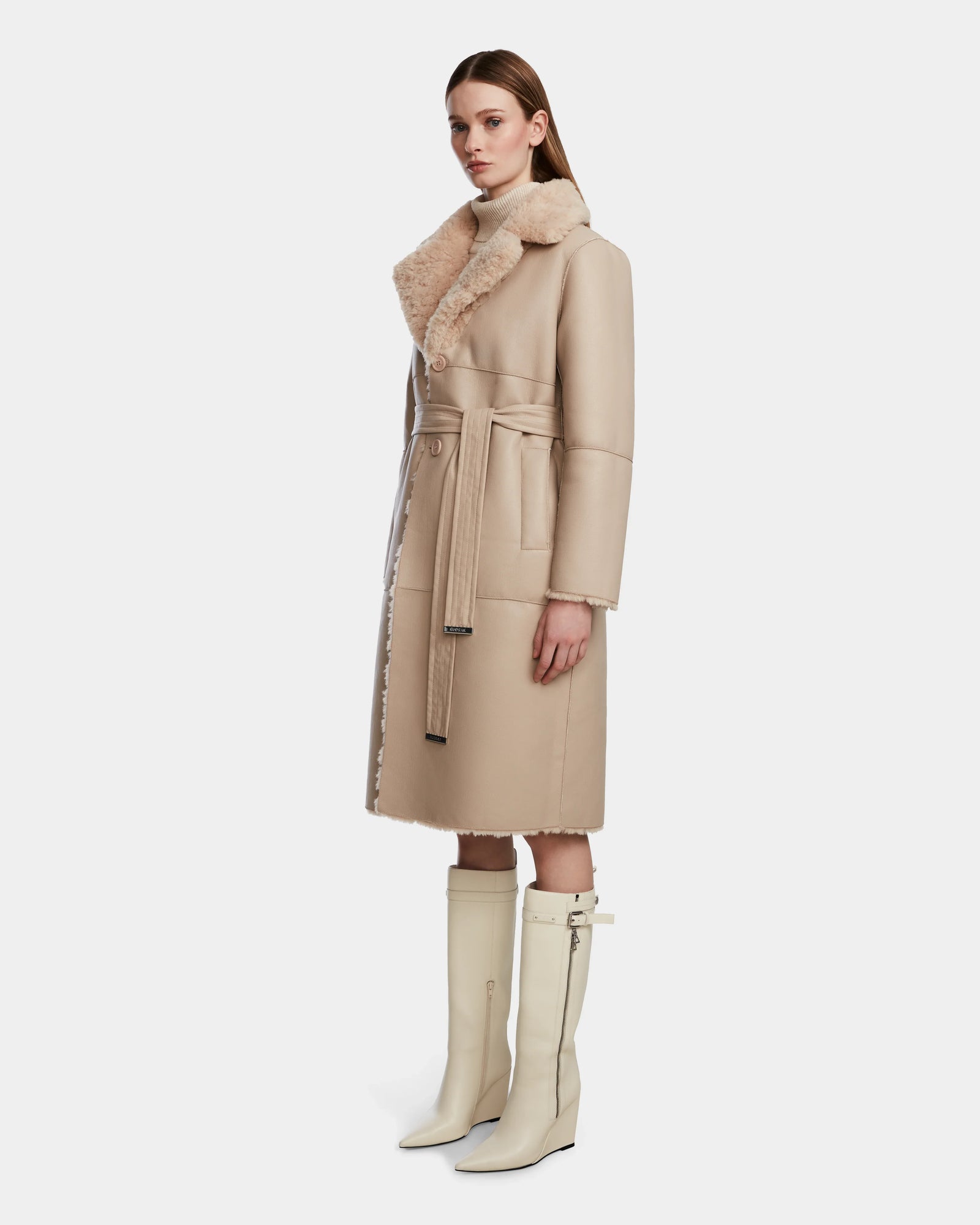 A woman wears the Rudsak ANNABELLE - L124544 Sand long vegan leather coat with a belt and wide collar, paired with knee-high cream boots, standing slightly turned against a plain white background.