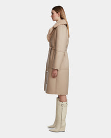A woman in profile wears the Rudsak ANNABELLE - L124544 Sand women's reversible teddy coat with a shearling collar and belt, paired with knee-high off-white boots. She has straight light brown hair and stands against a neutral background.