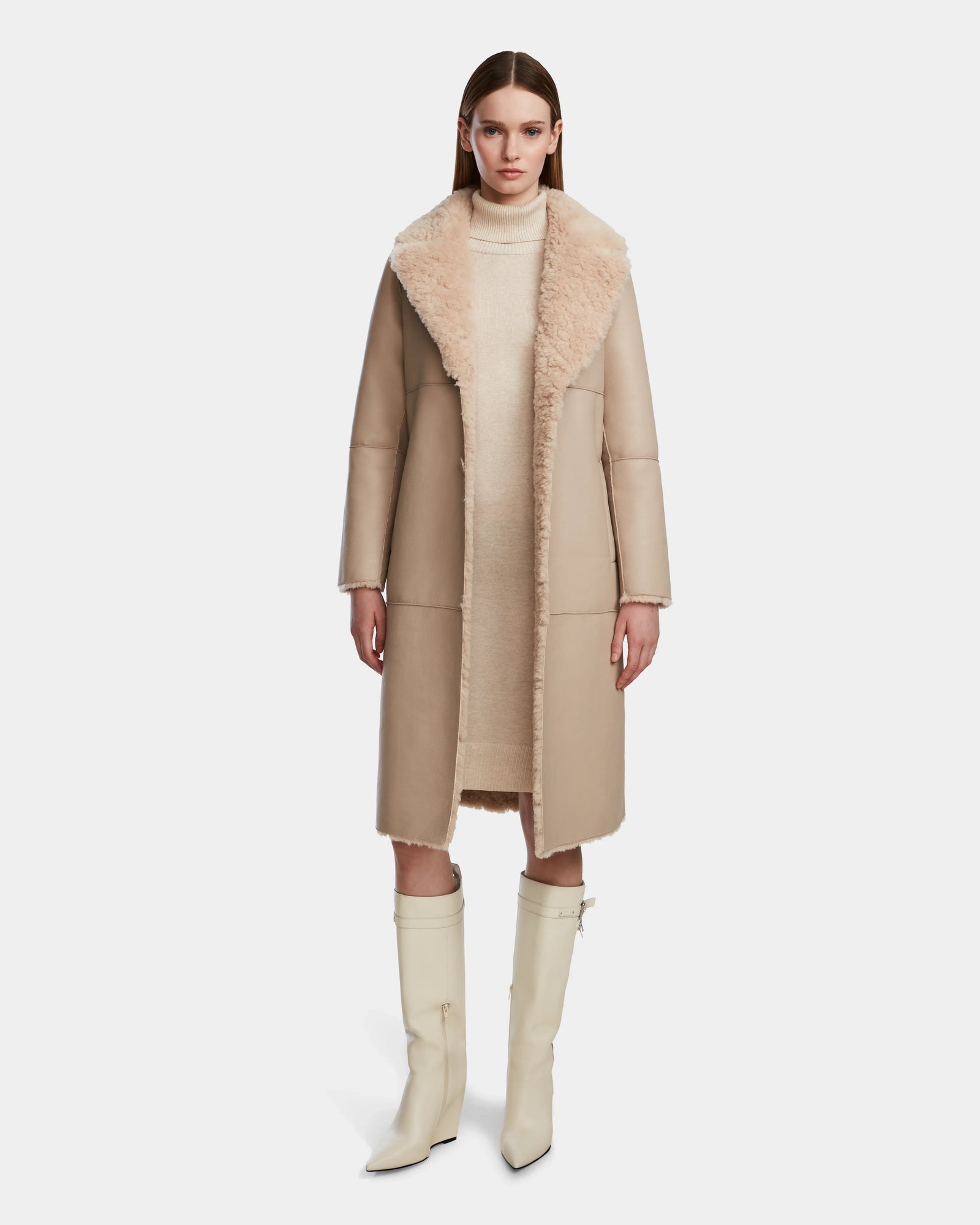 A woman faces forward, wearing the Rudsak ANNABELLE - L124544 Sand reversible teddy coat with a large fuzzy collar over a light knit dress and knee-high off-white boots. The background is plain white.