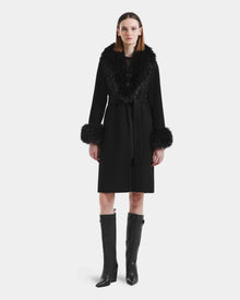 A woman stands facing forward in the Rudsak ARTIS - L125575 Black women’s wool coat with a belted waist, wide faux fur collar, fur-trimmed cuffs, and tall black heeled boots against a plain white background.
