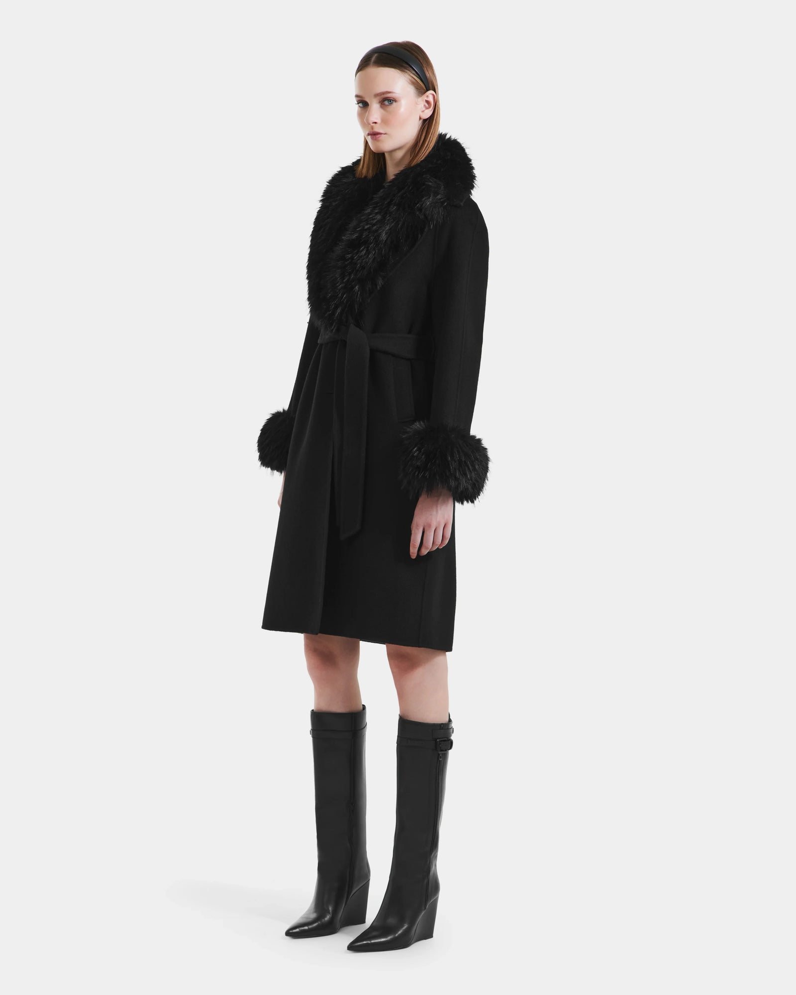 A woman with straight brown hair wears the Rudsak ARTIS - L125575 Black women’s wool coat featuring a faux fur collar and cuffs, belted at the waist, paired with black knee-high boots against a plain white background.