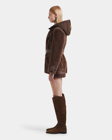 A woman with straight blonde hair faces left, modeling the Rudsak ASPEN F - 8125604 Mocha faux fur jacket with a removable down-insulated hood and tall brown suede boots, standing against a plain white background.