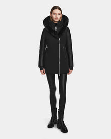 A woman stands facing forward against a plain white background, wearing the Rudsak AURELLE - L124538 Black coat with vegan leather sleeves and a large fur-trimmed collar, paired with black leggings and ankle boots.