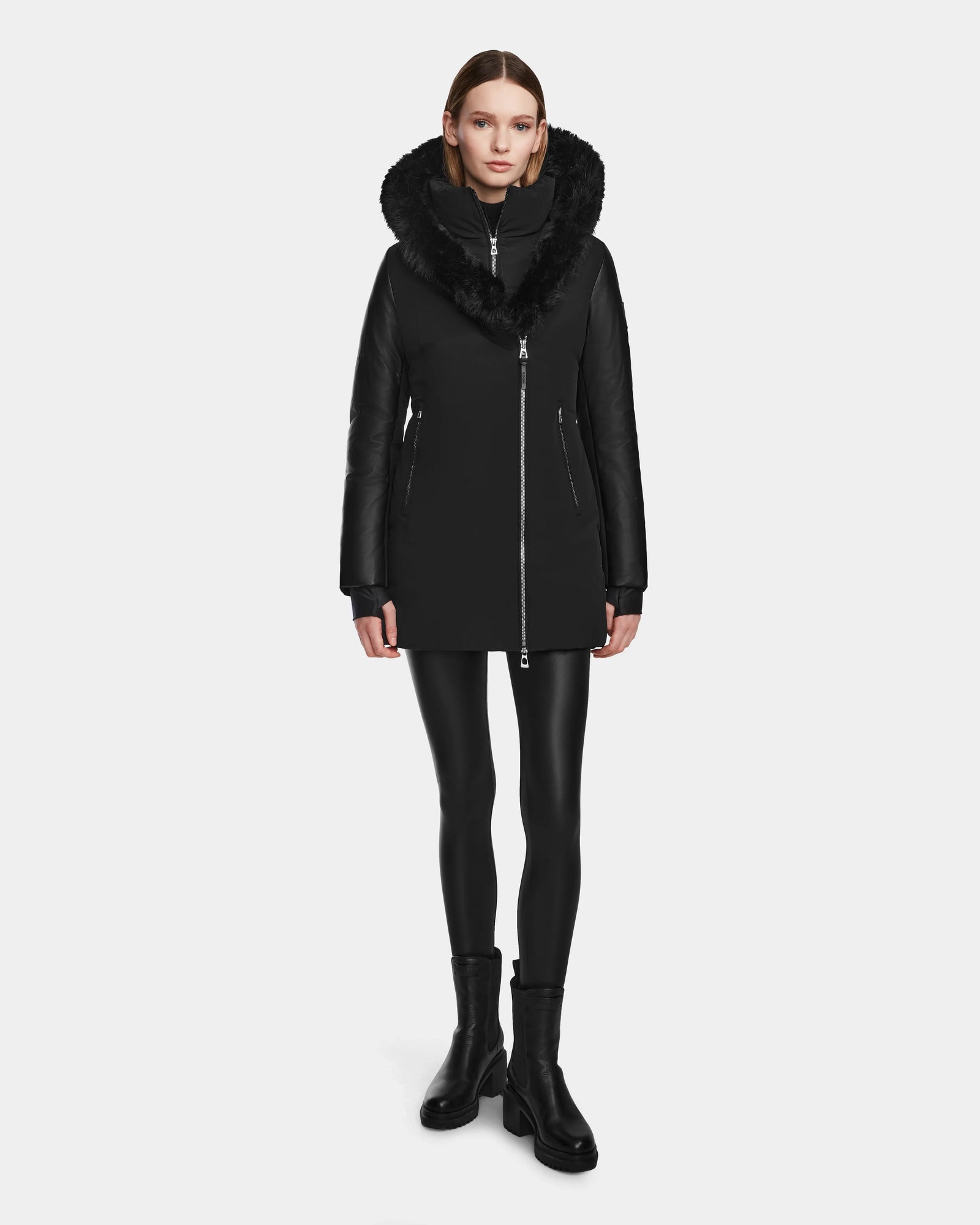 A woman stands facing forward against a plain white background, wearing the Rudsak AURELLE - L124538 Black coat with vegan leather sleeves and a large fur-trimmed collar, paired with black leggings and ankle boots.