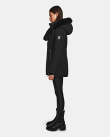 A woman wearing the Rudsak AURORE - L123561 Black/Black women's short fitted down parka with faux fur trim, paired with black leggings and boots, stands in profile against a plain white background.