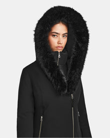 A woman wears the Rudsak AURORE - L123561 Black/Black women’s short fitted down parka with a faux fur-trimmed hood, zipped to her neck. The weather-resistant parka’s high loft down fill keeps her warm as she faces a plain background.