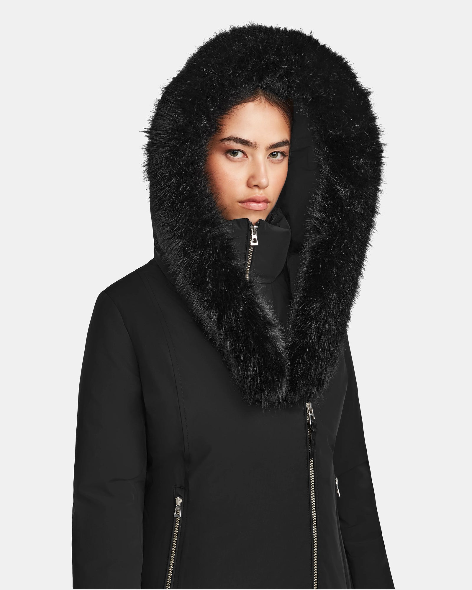 A woman wears the Rudsak AURORE - L123561 Black/Black women’s short fitted down parka with a faux fur-trimmed hood, zipped to her neck. The weather-resistant parka’s high loft down fill keeps her warm as she faces a plain background.