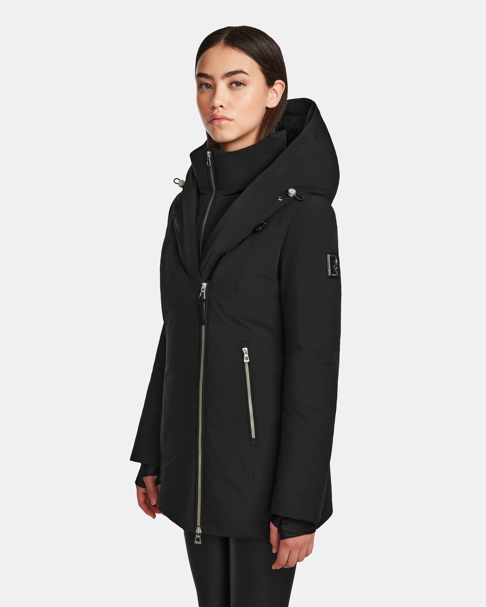A woman wearing the Rudsak AURORE - L123561 Black/Black short fitted down parka with an asymmetrical zipper and front zippered pockets stands slightly turned against a plain light background.