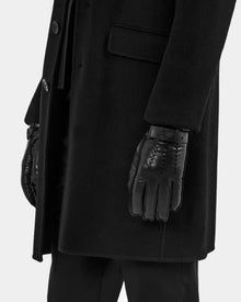 A person wears Rudsak's BORIS - M425001 Black coat, black pants, and men’s leather gloves with wool-cashmere lining, standing with their arm by their side against a plain light background—a true cold-weather essential.