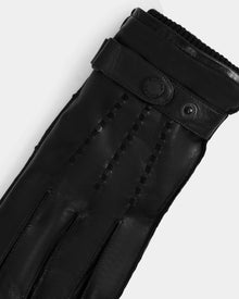 A close-up of Rudsak’s BORIS - M425001 Black men’s leather gloves, featuring decorative stitching, a snap button strap, ribbed wrist opening, and soft wool-cashmere lining—an essential for cold weather.