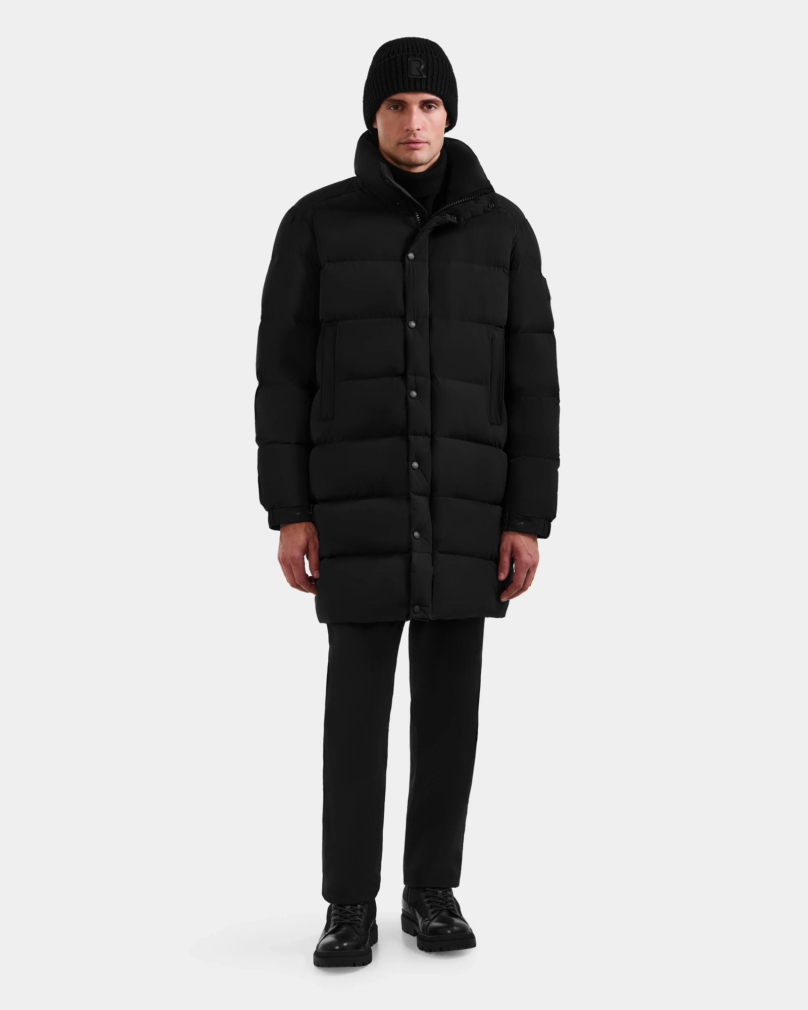 A person models the Rudsak BRAYDEN M-M - M125535 Black men’s down puffer coat, black pants, shoes, and a knit beanie against a white background, demonstrating winter style versatility.