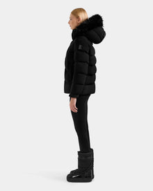 A person in profile stands against a light gray background, wearing the Rudsak BREE M - L125582 Black women’s down puffer with a removable fur-trimmed hood, paired with black leggings and winter boots—a must-have for cold weather.