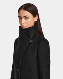 A woman with long, straight brown hair wears the Rudsak CAROLINE - L124543 Black, a high-collared, knee-length premium wool-blend coat, buttoned up and looking slightly to the side against a plain light background.