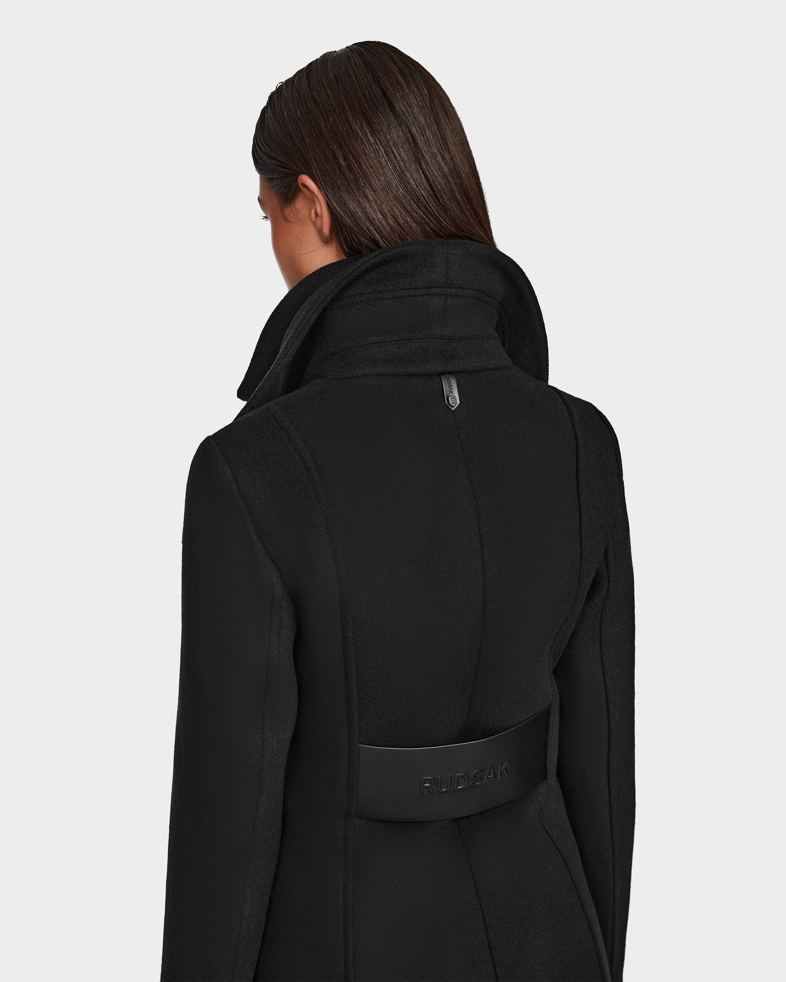 A woman with straight brown hair is seen from the back wearing the Rudsak CAROLINE - L124543 Black knee-length coat with a high collar and a branded black strap, set against a plain light gray background.