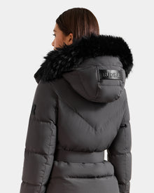 A woman with straight brown hair wears the Rudsak CHRISTELLE M - L125542 Grey, a weather-resistant quilted down puffer with a black faux fur-trimmed hood. 
