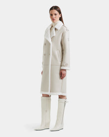 A woman wears the Rudsak DARCY - L125539 Vanilla long winter coat with white fur trim and matching knee-high boots, facing forward with a neutral expression against a plain white background.
