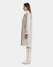 A woman stands in profile wearing the Rudsak DARCY - L125539 Vanilla long light beige women's winter coat with white faux sherpa teddy lining, a white turtleneck, and knee-high white zippered boots against a plain white background.