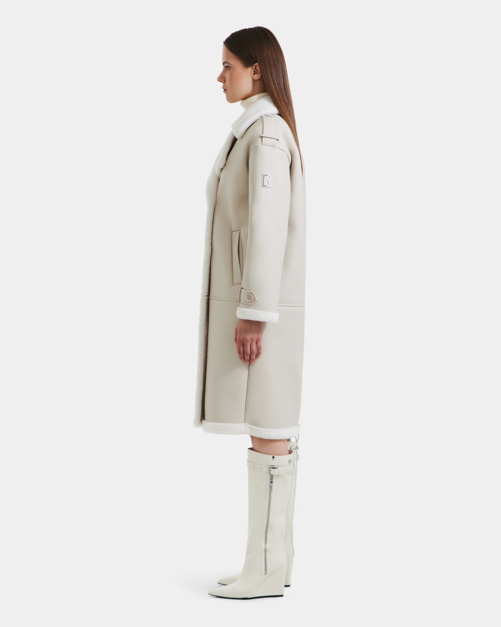 A woman stands in profile wearing the Rudsak DARCY - L125539 Vanilla long light beige women's winter coat with white faux sherpa teddy lining, a white turtleneck, and knee-high white zippered boots against a plain white background.