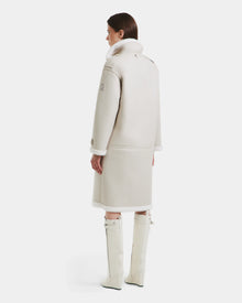 A woman stands facing away in the DARCY - L125539 Vanilla winter coat by Rudsak, its elegant faux sherpa teddy texture and high collar highlighted against a white background, paired with matching knee-high boots.