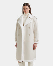 A woman stands facing forward, wearing the Rudsak DARCY - L125539 Vanilla long faux sherpa coat with wide lapels and double-breasted buttons over a light turtleneck, set against a plain white background.