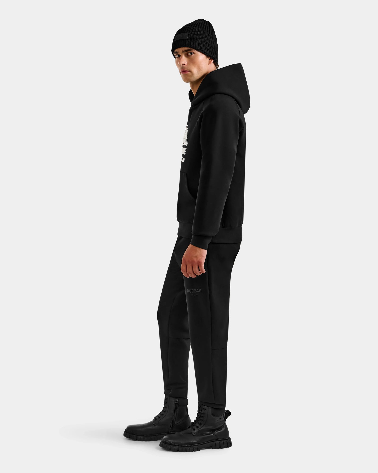 A person wearing the Rudsak DARWIN - M525502 Black hoodie, black beanie, pants, and boots stands sideways against a plain white background.