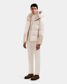 A man faces forward against a plain background, wearing Rudsak's DAVID - M125508 Vanilla men’s shearling puffer jacket with a hood, styled with a white turtleneck, cream pants, and brown suede boots.