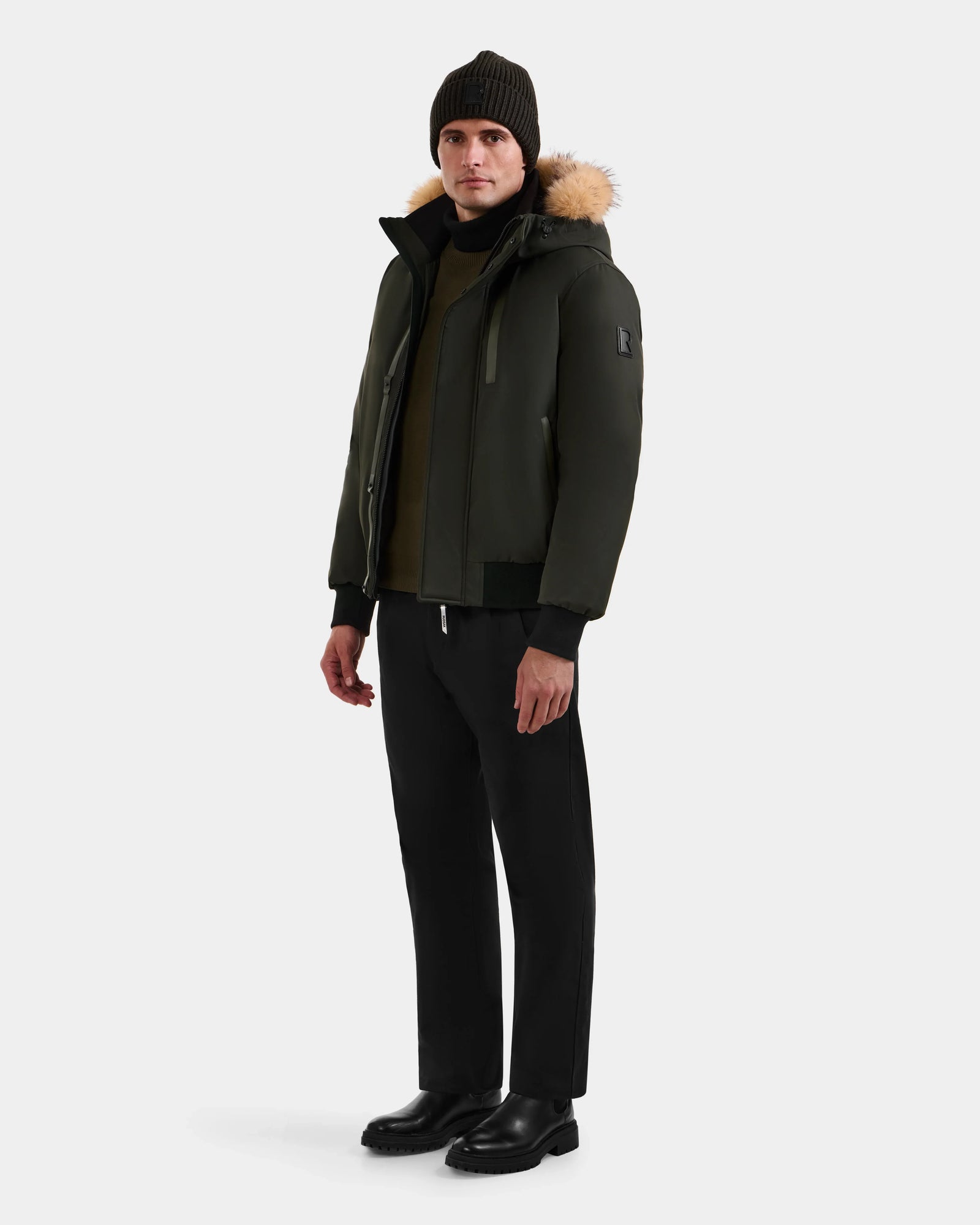 A man wears the Rudsak DEACON - M123513 Dark Olive men's winter coat with a fur-trimmed hood, paired with black pants, boots, and a dark knit beanie against a plain light gray background.