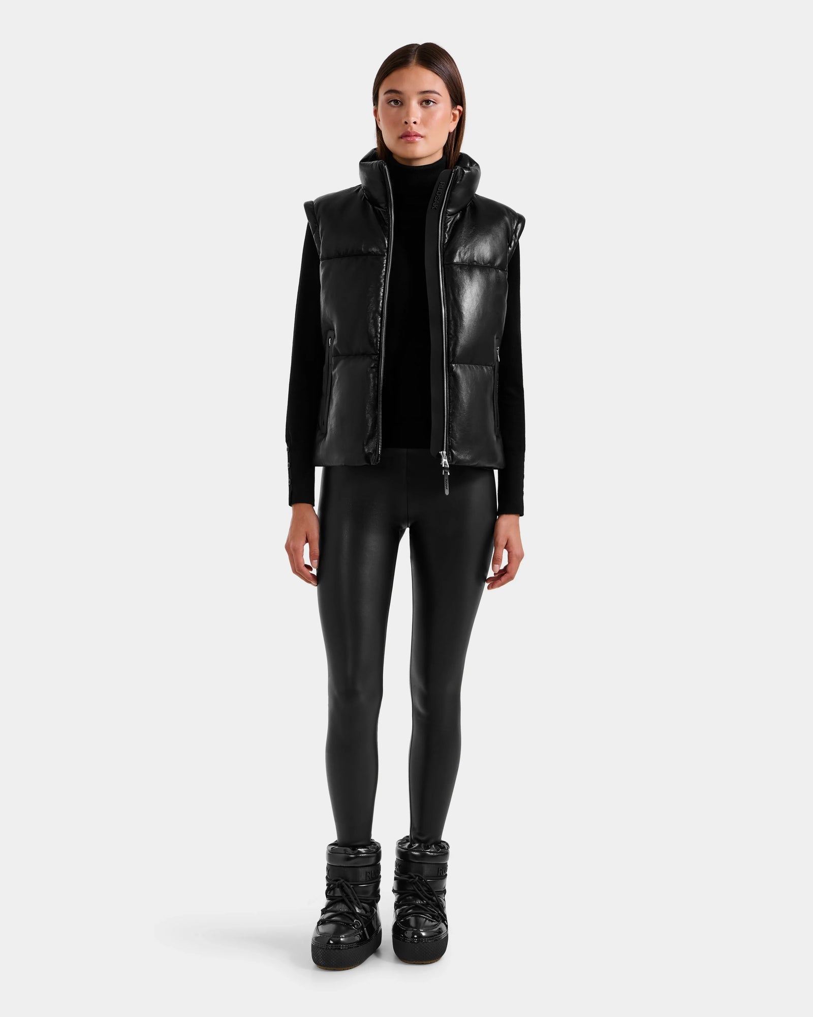 A woman faces forward wearing the Rudsak DELILAH PX - L125537 Black down vest, paired with a black turtleneck, faux leather leggings, and puffer boots against a plain light gray background.