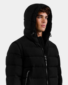 A man in the Rudsak DEMIAN M-R - M123504 Black men's down puffer jacket stands against a plain light background, hood up and looking to the side with a neutral expression.