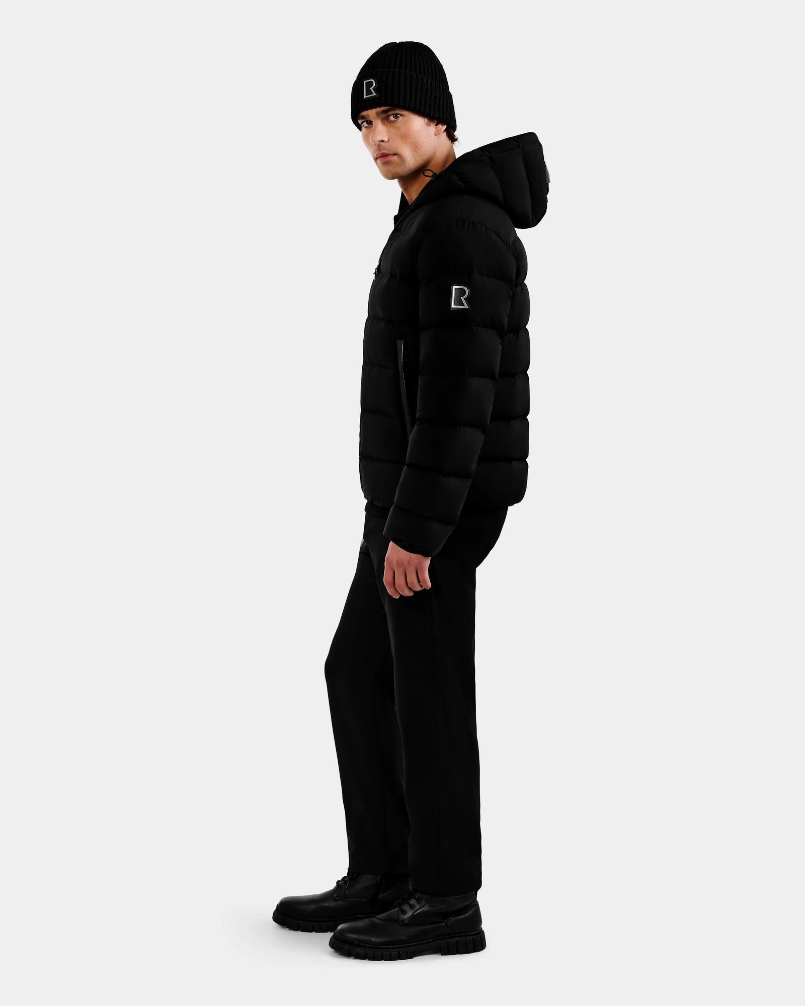 A person stands sideways in all black, wearing the Rudsak DEMIAN M - M123507 down puffer and a knit beanie, both with matching "R" patches. The jacket is made from recycled fabric. The background is plain light gray.