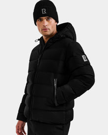 A man wears the DEMIAN M - M123507 Black down puffer jacket with hood and a black knit beanie, both by Rudsak and featuring the R logo. Made from sustainable recycled fabric, he stands against a light gray background facing forward.