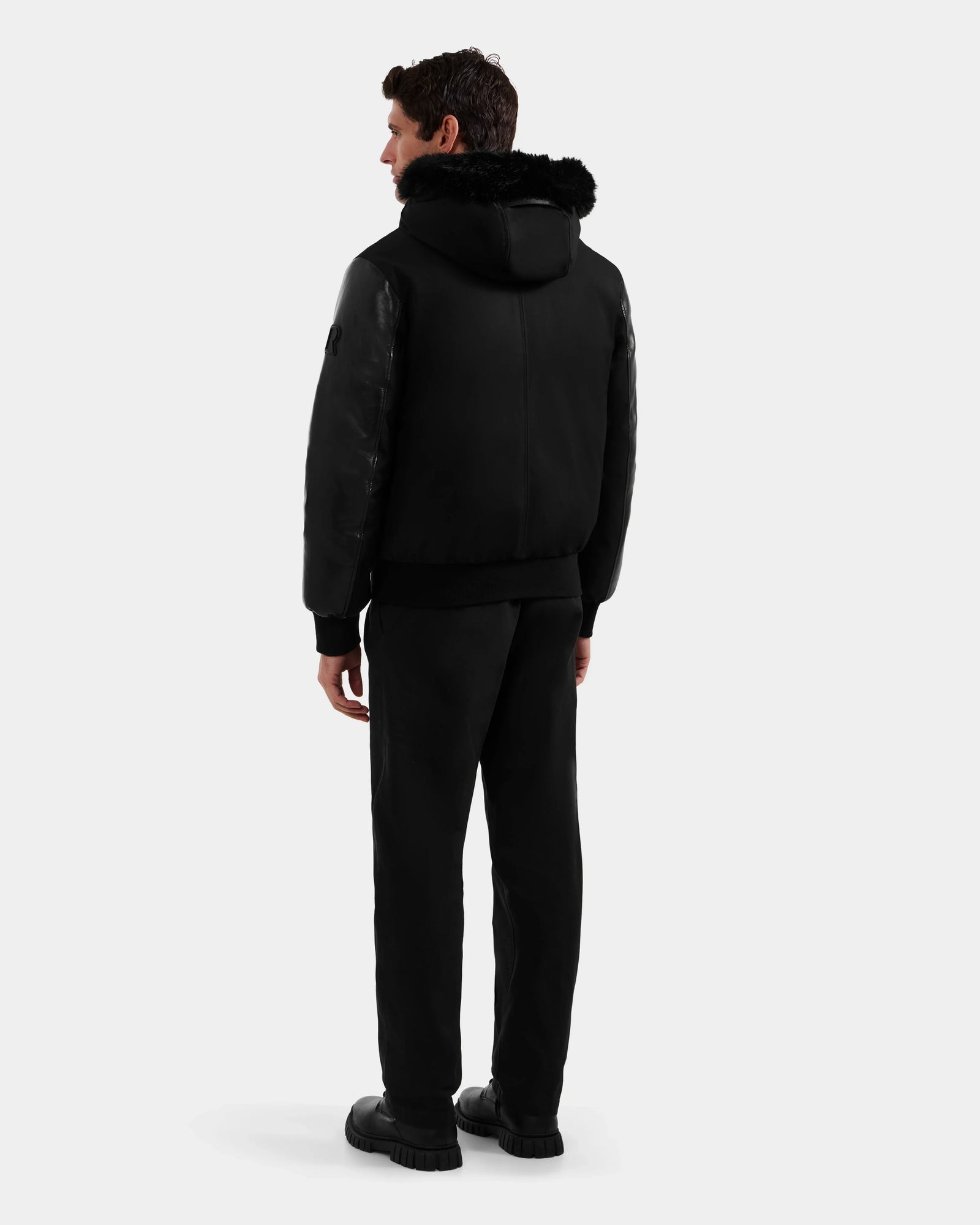 A man faces away wearing the Rudsak DERON - M125539 Black men’s down bomber with vegan-leather sleeves, paired with black pants and shoes, set against a plain light gray background.