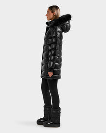 A woman stands in profile wearing the Rudsak ELISA S - L125572 Black quilted puffer jacket with a black fur-lined hood, paired with black leggings and winter boots, against a plain light gray background.