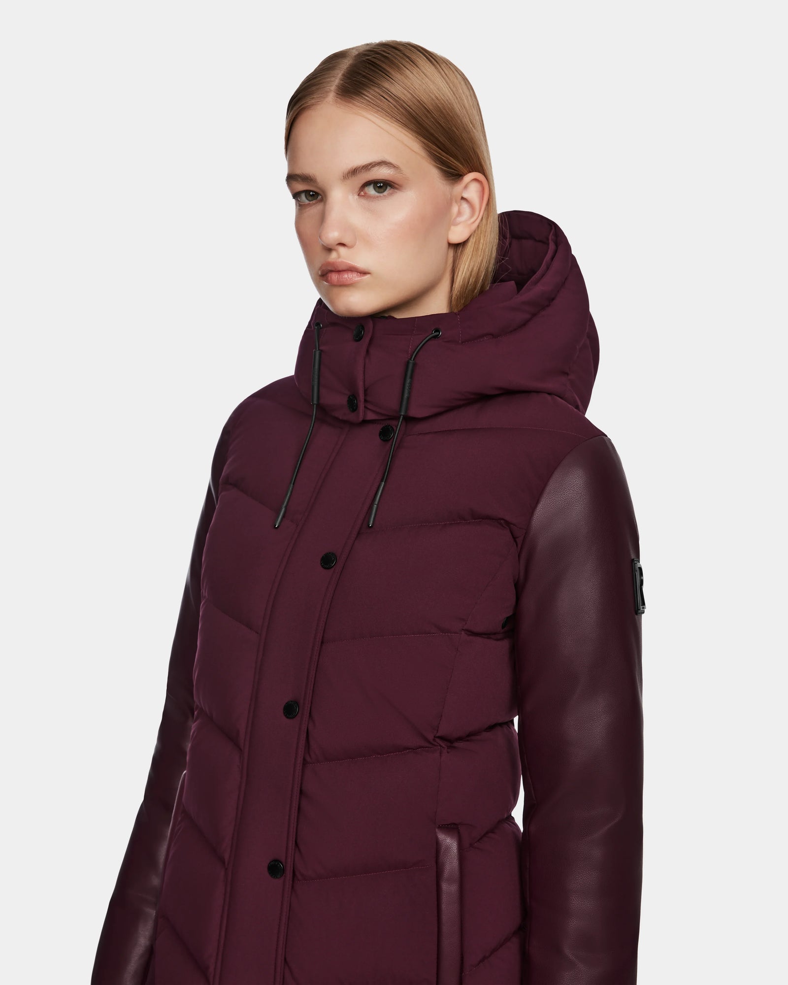 A woman with straight blonde hair wears the Rudsak ELSA - L123535 Wine women’s quilted down puffer jacket with leather sleeves and a hood, standing against a plain light background.