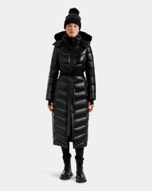 A person stands against a plain light background wearing the Rudsak ELSIE S - L124536 Black women’s shiny black, water-resistant maxi down puffer with fur-trimmed hood, paired with a black beanie with pom-pom, black boots, and pants.