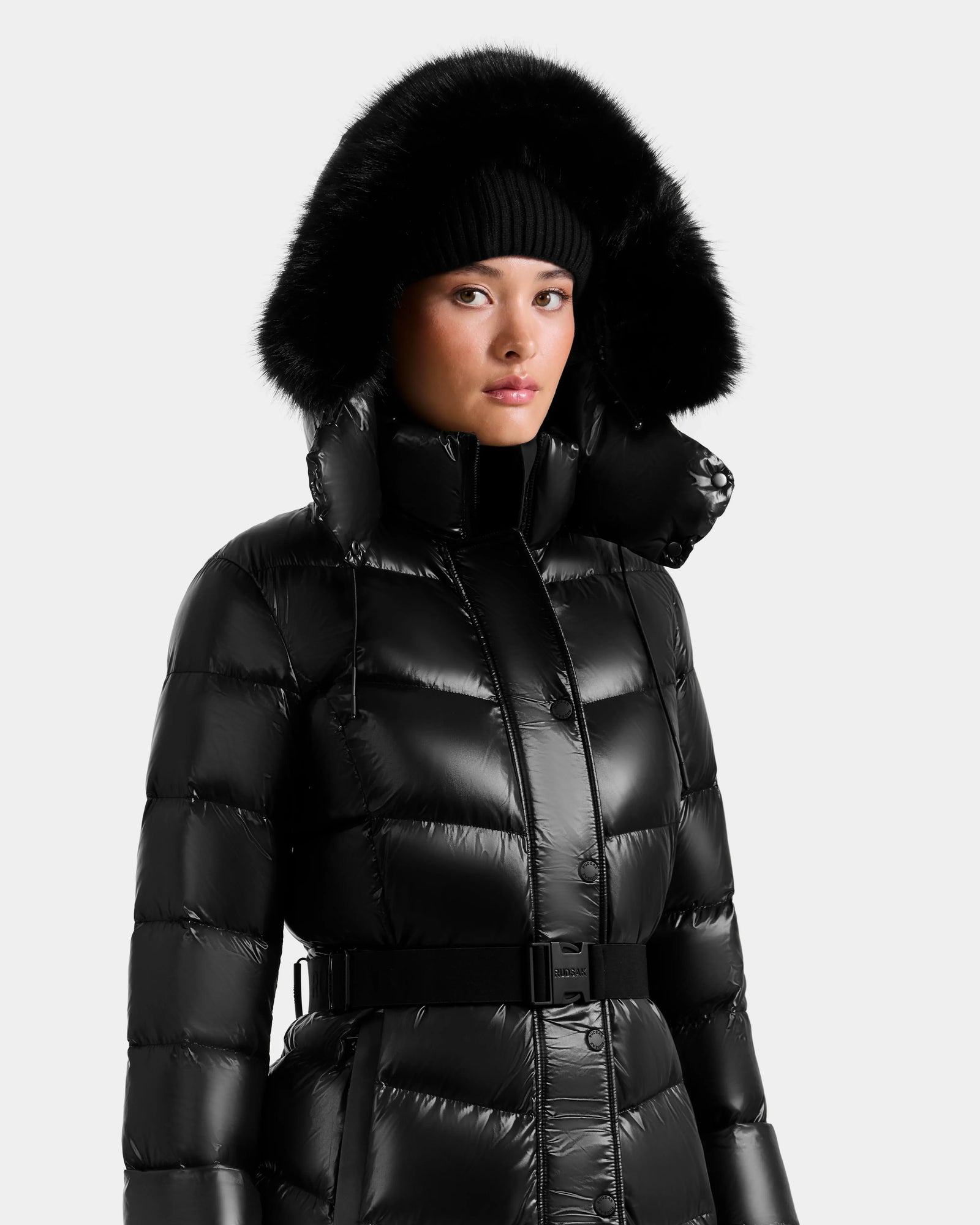A person wears the Rudsak ELSIE S - L124536 Black women’s maxi puffer coat with a belted waist, high collar, and black fur-trimmed hood, paired with a black knit beanie against a plain, light background.