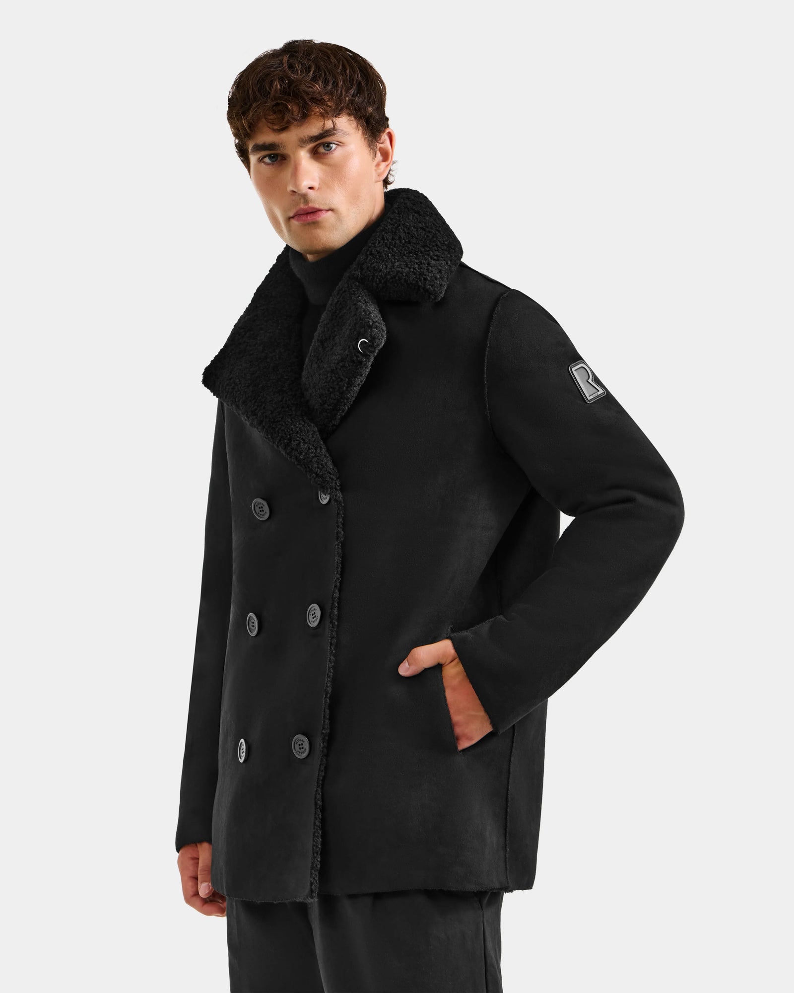A man with short curly hair wears the Rudsak ELTON - M125534 Black reversible overcoat with a large faux shearling collar and double-breasted buttons, standing against a light background and looking at the camera, hand in pocket.