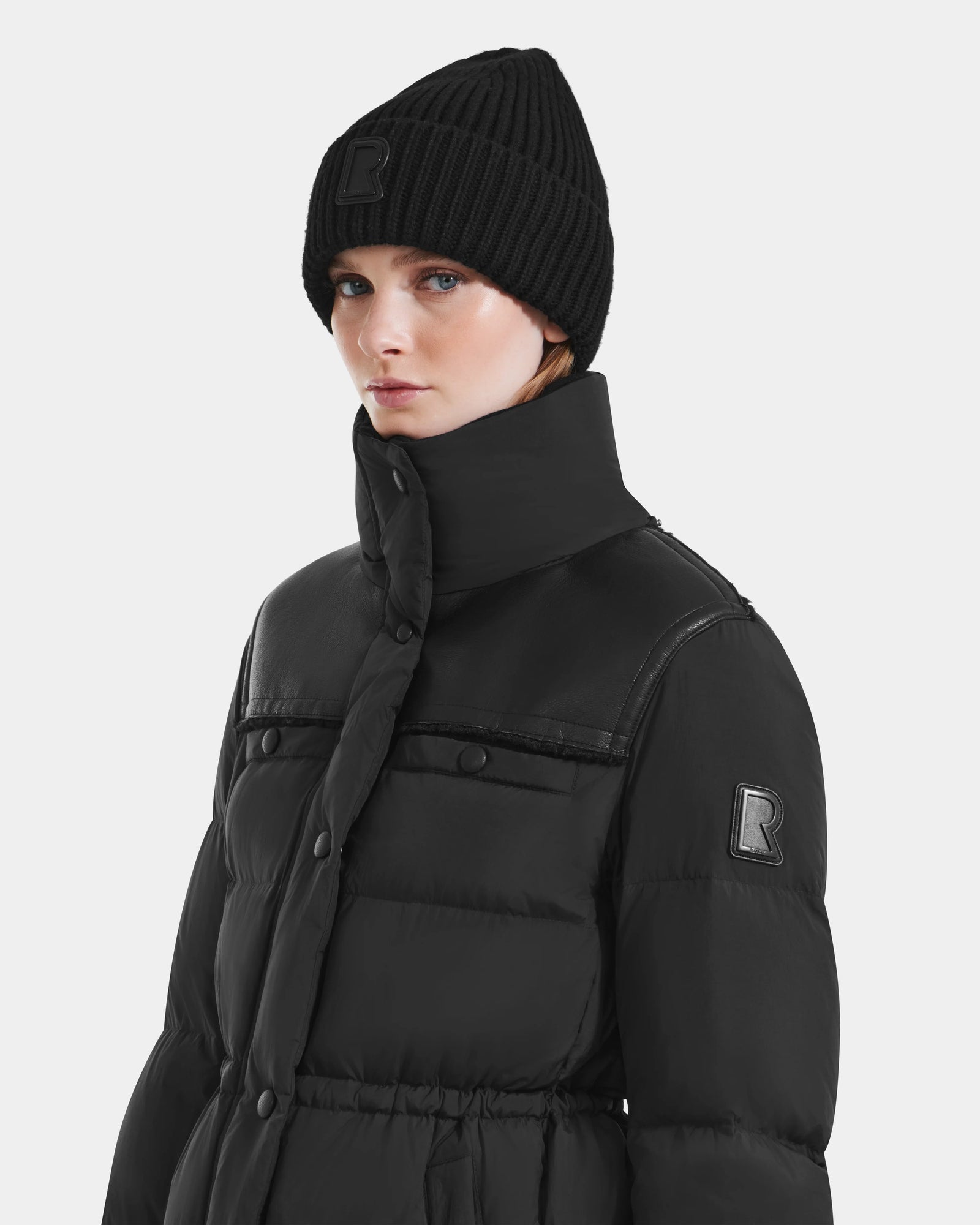A person wears the EMBER M-M - L125524 Black puffer jacket by Rudsak, with vegan-leather piping, high collar, and raised R logo, paired with a black beanie. The background is plain and light-colored.