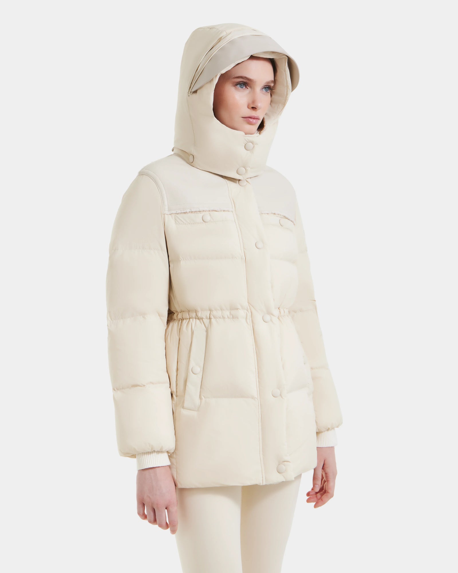 A woman wears the Rudsak EMBER M-M - L125524 Vanilla vegan-leather puffer jacket with a hood, button and zipper closure, and light-colored pants, standing against a plain white background.