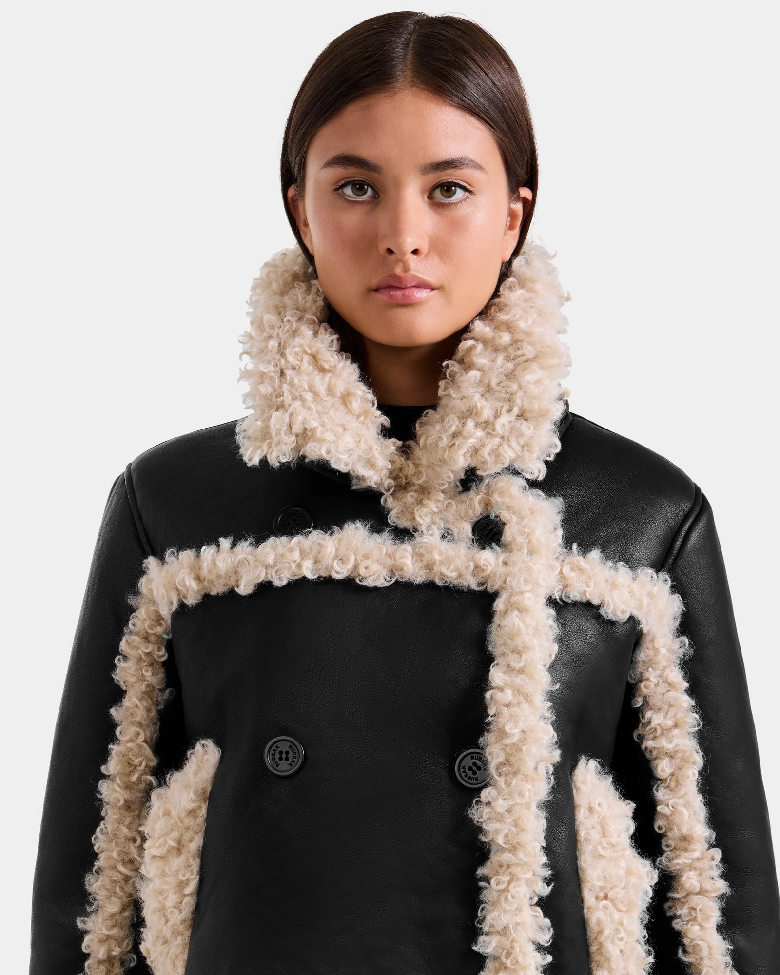 A woman with straight brown hair wears the Rudsak EMERY - L125548 Black vegan-leather jacket with cream faux shearling trim and large buttons, looking directly at the camera against a plain light background.
