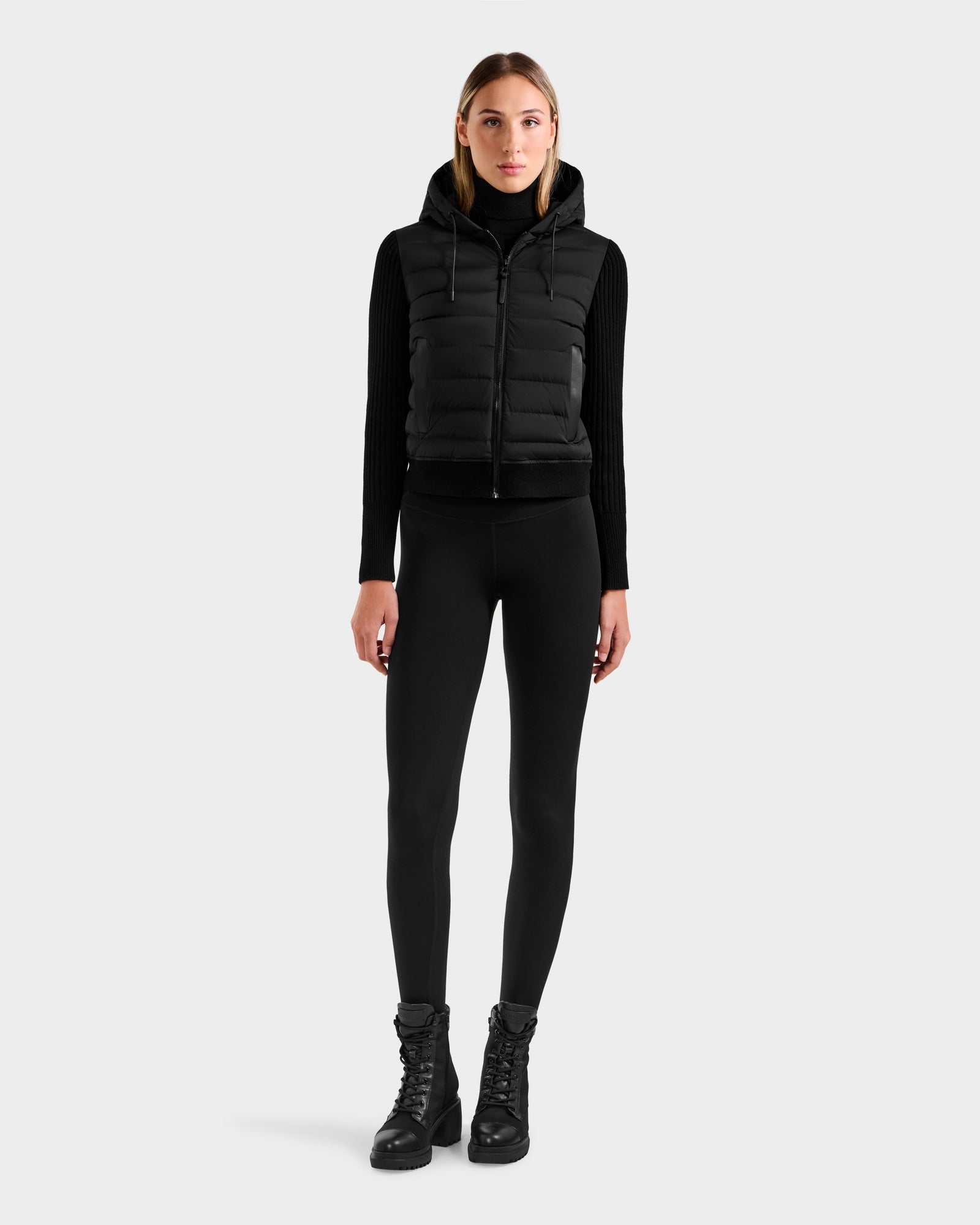 A woman faces forward against a light gray background, wearing the Rudsak EMILY - L125515 Black women’s hooded down bomber vest over a black long-sleeve top, black leggings, and black lace-up boots.