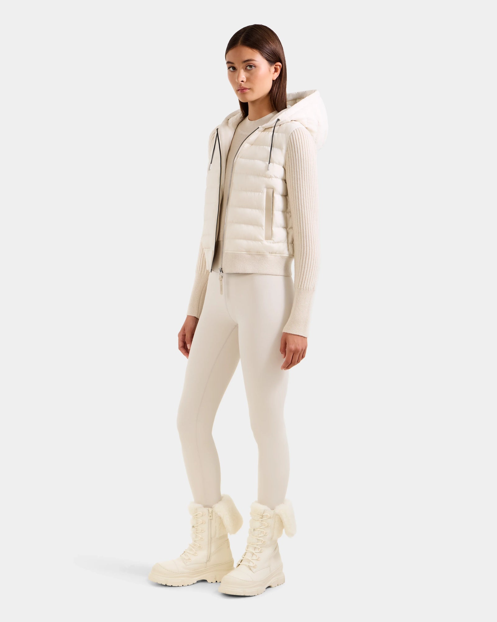 A woman stands in a neutral pose wearing the Rudsak EMILY - L125515 Vanilla off-white hooded down bomber jacket with matching fitted leggings and white lace-up winter boots, against a plain light background.