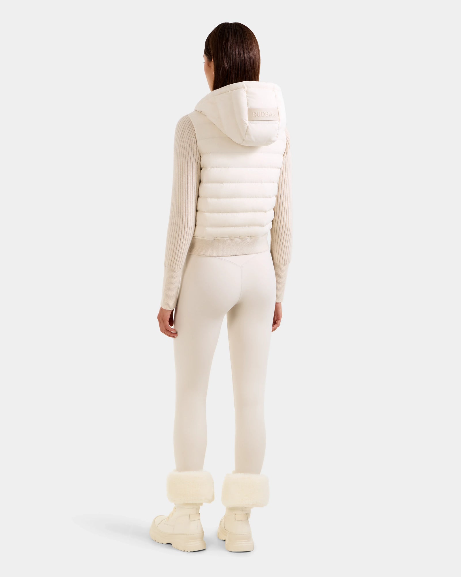 A person faces away, wearing the Rudsak EMILY - L125515 Vanilla women’s hooded down bomber with ribbed sleeves, paired with fitted leggings and boots with fluffy cuffs, set against a plain light background.