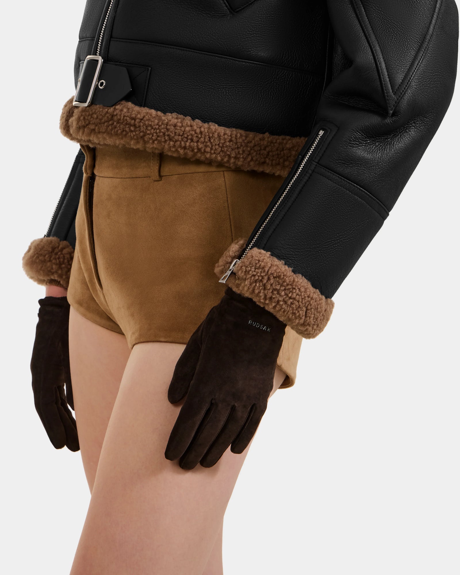 A person wears Rudsak’s FLORA - L425542 Espresso leather jacket with brown shearling trim, paired with tan suede shorts and women's suede gloves. The image highlights the chic winter look from waist to mid-thigh.