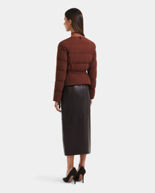 A woman faces away in the Rudsak FRANCESCA - 8125021 Rust puffer jacket, paired with a black midi leather skirt and black high heels, set against a plain white background.