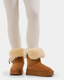 A person wears white leggings and Rudsak’s INESA - L225026 Tan women’s suede platform boots with thick, cream-colored shearling cuffs and cozy wool lining, standing on a white background.
