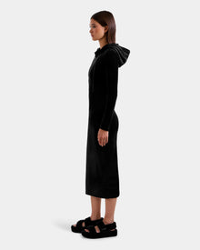 A woman stands in profile facing left, wearing the Rudsak ISABELLA - L525502 Black velour hooded midi dress with long sleeves, paired with black platform sandals against a plain light background.